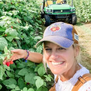 Brooke in a hat showing a raspberry bush with ripe berries
